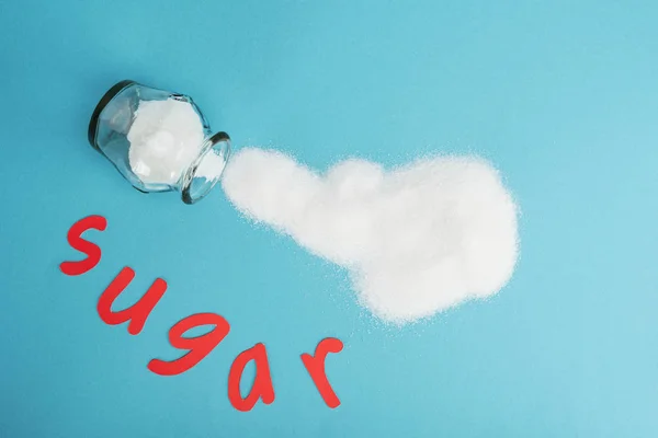 Vista dall'alto di carta rossa taglio parola zucchero vicino vaso di vetro e cristalli di zucchero spruzzato su sfondo blu — Foto stock