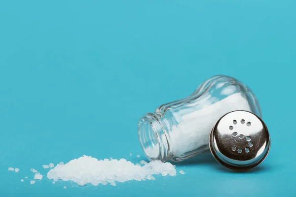 Open salt shaker and metallic cap near sprinkled salt on blue background with copy space — Stock Photo