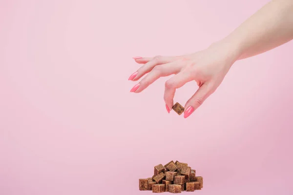 Vue partielle de la femme tenant un cube de sucre près d'un tas de cubes de sucre brun sur fond rose — Photo de stock
