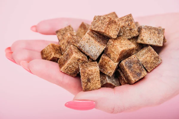 Cropped view of woman with handful of brown sugar cubes on pink background — Stock Photo