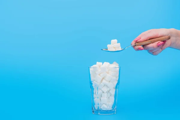 Vista ritagliata di donna che tiene cucchiaino da tè con zucchero vicino a vetro con cubetti di zucchero isolati su blu — Foto stock