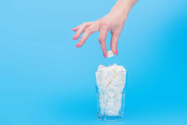 Vista parcial de la mujer sosteniendo cubo de azúcar cerca de vidrio lleno de cubos de azúcar blanco aislado en azul - foto de stock