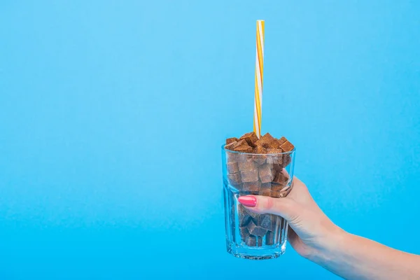 Abgeschnittene Ansicht einer Frau mit Glas mit Stroh und braunen Zuckerwürfeln isoliert auf blauem Grund — Stockfoto