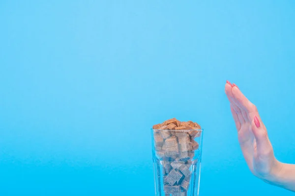 Vista recortada de la mujer que muestra gesto de basura cerca de vidrio con cubos de azúcar moreno aislado en azul — Stock Photo