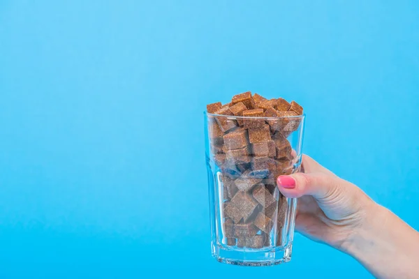 Частковий вигляд жіночої руки зі склянкою, повним коричневих кубиків цукру ізольовані на синьому — стокове фото