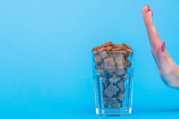 Vista parcial de la mujer que no muestra ningún gesto cerca de vidrio con cubos de azúcar moreno sobre fondo azul con espacio de copia - foto de stock