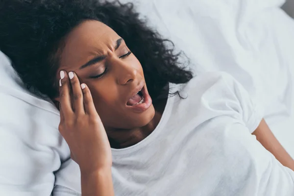 Mulher afro-americana infeliz deitada na cama e sofrendo de dor de cabeça — Fotografia de Stock