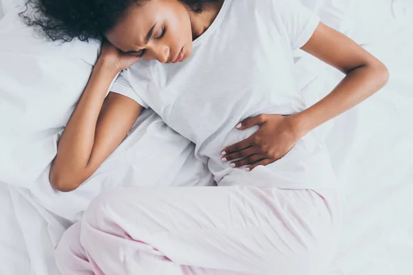 Pretty african american woman suffering from stomach pain while lying in bed — Stock Photo