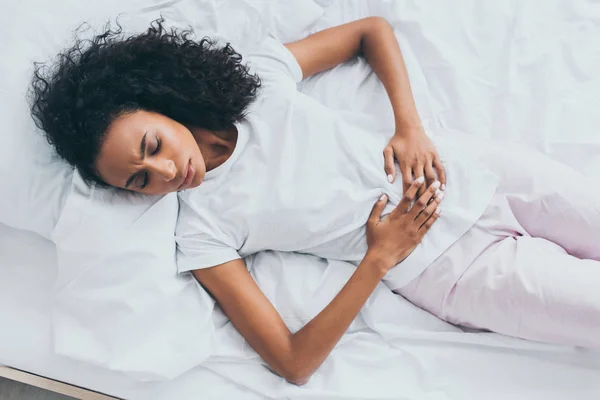 Beautiful african american woman suffering from abdominal pain while lying on white bedding — Stock Photo