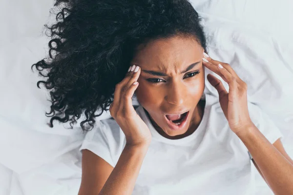 Mujer afroamericana agotada gritando mientras sufre de dolor de cabeza en la cama - foto de stock