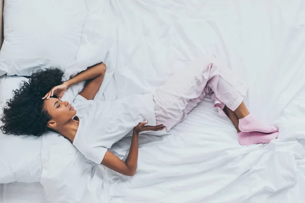 Vista superior de la infeliz mujer afroamericana que sufre de dolor de espalda mientras está acostada sobre ropa de cama blanca - foto de stock