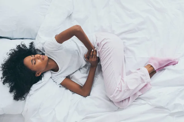 Vue de dessus de jolie femme afro-américaine souffrant de douleurs abdominales alors qu'elle était allongée sur une literie blanche avec les yeux fermés — Photo de stock