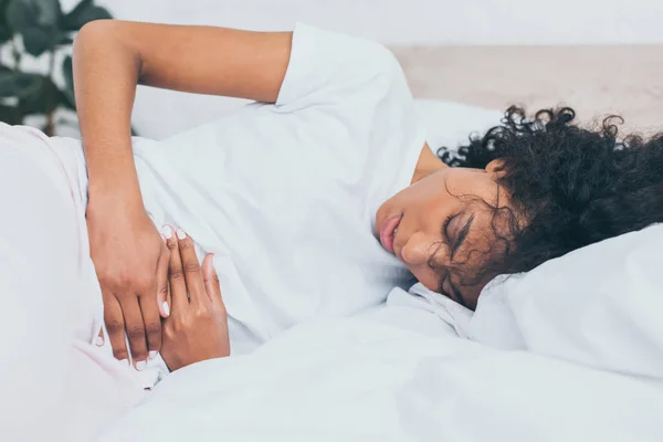 Exhausted african american woman suffering from stomach pain while lying in bed — Stock Photo