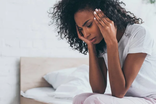 Pretty african american woman holding hands near head while suffering from headache in bedroom — Stock Photo