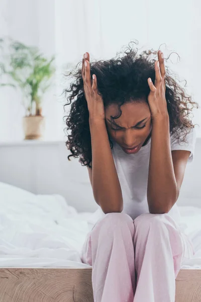 Unglückliche afrikanisch-amerikanische Frau hält Hände in Kopfnähe, während sie unter Kopfschmerzen im Schlafzimmer leidet — Stockfoto