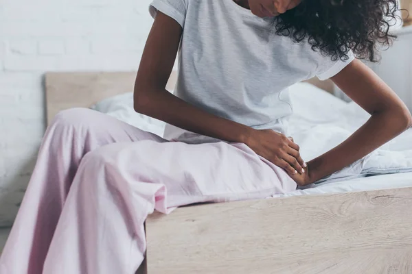 Abgeschnittene Ansicht einer Afroamerikanerin, die unter Schmerzen in der Hüfte leidet, während sie auf dem Bett sitzt — Stockfoto