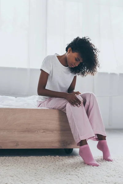 Mulher americana africana bonita que sofre de dor no joelho enquanto sentado na cama — Fotografia de Stock