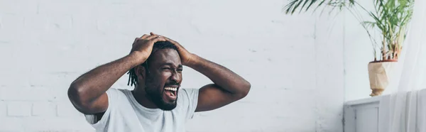 Tiro panorâmico de homem afro-americano gritando enquanto sofre de dor de cabeça no quarto — Fotografia de Stock