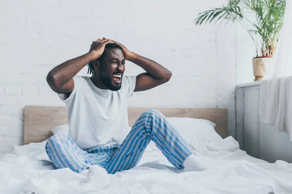 Hombre afroamericano gritando mientras sufre de dolor de cabeza en el dormitorio - foto de stock