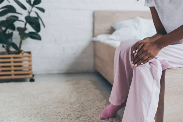 Abgeschnittene Ansicht einer afrikanisch-amerikanischen Frau, die unter Knieschmerzen leidet, während sie auf dem Bett sitzt — Stockfoto
