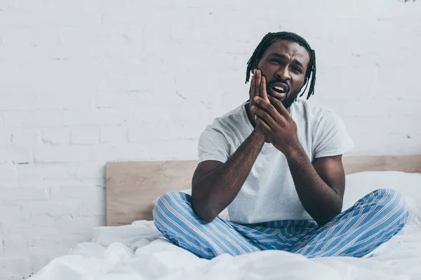 Handsome african american man in pajamas suffering from tooth pain in bedroom — Stock Photo