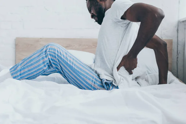 Giovane uomo afro-americano che soffre di mal di schiena mentre è seduto su biancheria da letto — Foto stock
