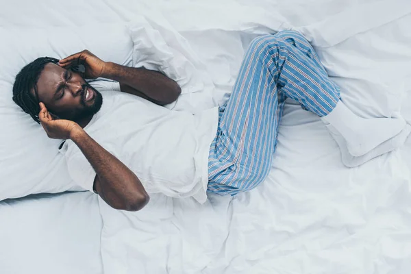 Vista superior do homem americano africano exausto que sofre de dor de cabeça enquanto deitado em roupa de cama branca — Fotografia de Stock