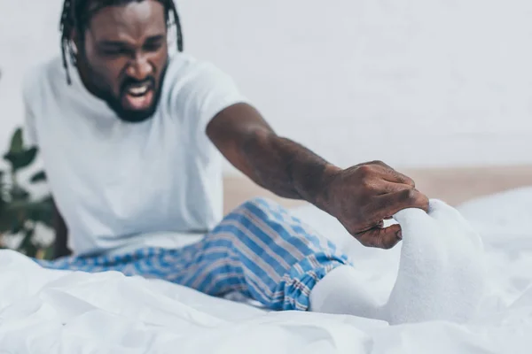 Selektiver Fokus des schreienden afrikanisch-amerikanischen Mannes, der unter Fußschmerzen im Schlafzimmer leidet — Stockfoto