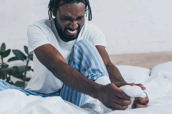 Junger afrikanisch-amerikanischer Mann leidet unter Fußschmerzen, während er auf Bettwäsche sitzt — Stockfoto