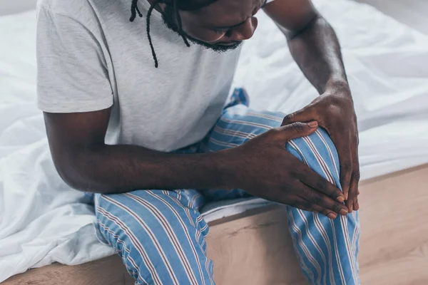 Vista ritagliata di giovane africano che soffre di dolore al ginocchio in camera da letto — Foto stock