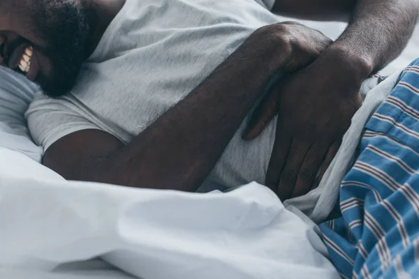 Vista parcial del hombre afroamericano que sufre de dolor abdominal en el dormitorio - foto de stock
