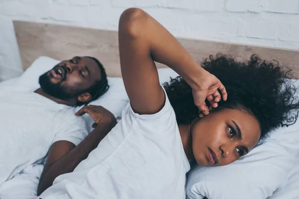 Селективный фокус африканской американки, затыкающей уши пальцем, когда она лежит рядом с храпящим мужем — стоковое фото