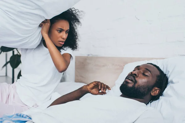 Angry african american woman holding pillow while sitting in bed near snoring husband — Stock Photo