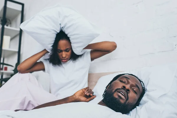 Unhappy african american woman covering head with pillow while sitting in bed near snoring husband — Stock Photo