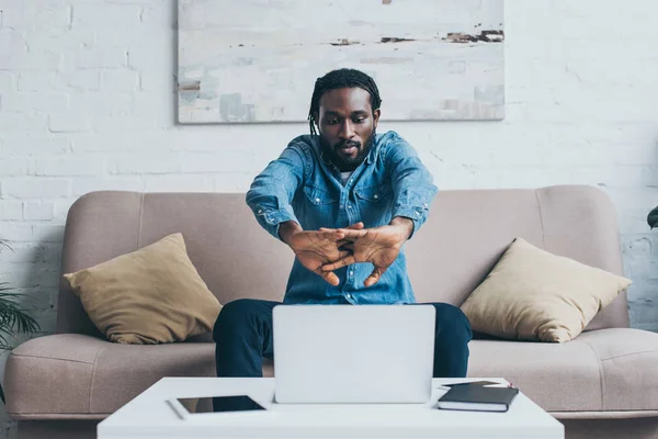 Bonito afro-americano homem sentado no sofá perto da mesa com dispositivos digitais — Fotografia de Stock