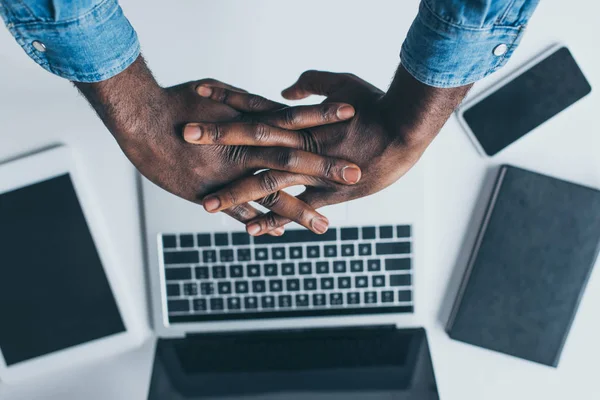 Vista recortada del hombre afroamericano sosteniendo las manos apretadas por encima de la mesa con computadora portátil, tableta digital y portátil - foto de stock