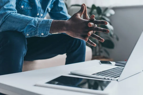 Vue recadrée de l'homme afro-américain assis avec les mains serrées près de la table avec ordinateur portable et tablette numérique — Photo de stock