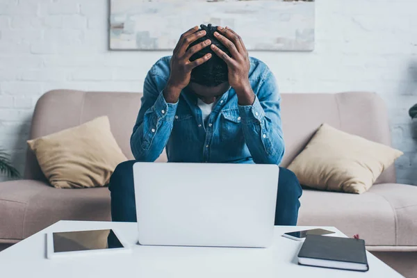 Homme afro-américain fatigué souffrant de maux de tête tout en étant assis près de la table avec des gadgets — Photo de stock