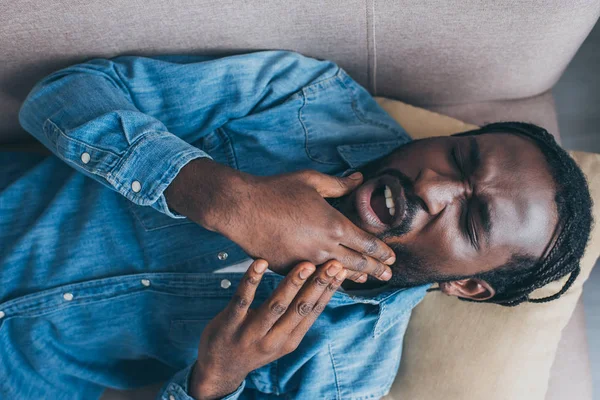 Vista superior del hombre afroamericano acostado en el sofá y que sufre de dolor en la mandíbula - foto de stock