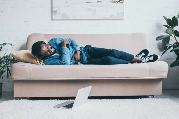 Young african american man suffering from stomach pain while lying on sofa near laptop on floor — Stock Photo