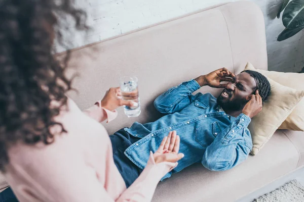 Mujer afroamericana sosteniendo un vaso de agua y pastillas mientras está de pie cerca del hombre que sufre de dolor de cabeza en el sofá — Stock Photo