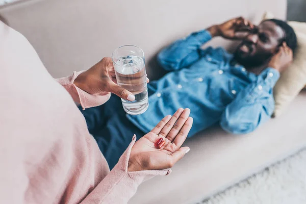 Vue recadrée de la femme afro-américaine tenant un verre d'eau et des pilules tout en se tenant près de l'homme souffrant de maux de tête sur le canapé — Photo de stock