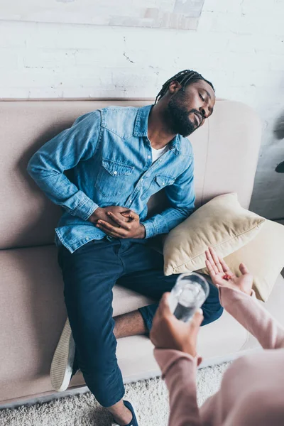 Visão parcial da mulher afro-americana dando copo de água e pílulas para o homem sentado no sofá e sofrendo de dor abdominal — Fotografia de Stock