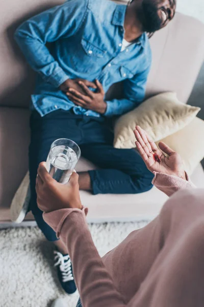 Vue recadrée de la femme afro-américaine donnant un verre d'eau et des pilules à l'homme assis sur le canapé et souffrant de douleurs abdominales — Photo de stock