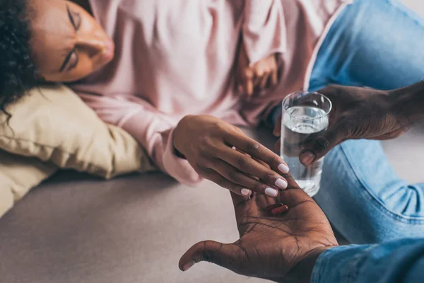 Частичный вид африканского американца, дающего стакан воды и таблетки женщине, лежащей на диване и страдающей от боли — стоковое фото