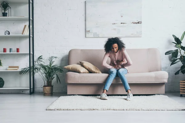 Young african american woman suffering from abdominal pain while sitting on sofa in living room — Stock Photo