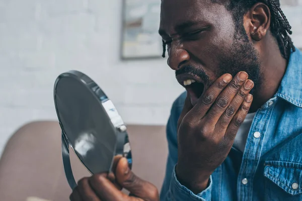 Aufgebrachter Afroamerikaner blickt in den Spiegel, während er unter Zahnschmerzen leidet — Stockfoto