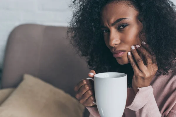 Unhappy african american woman looking at camera while suffering from tooth pain and holding coffee cup — Stock Photo