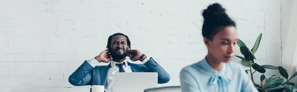 Colpo panoramico di manager afro-americano che soffre di mal di testa mentre è seduto in ufficio vicino a bella donna d'affari — Foto stock