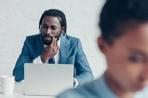 Unhappy african american businessman suffering from tooth pain while sitting in office near pretty colleague — Stock Photo